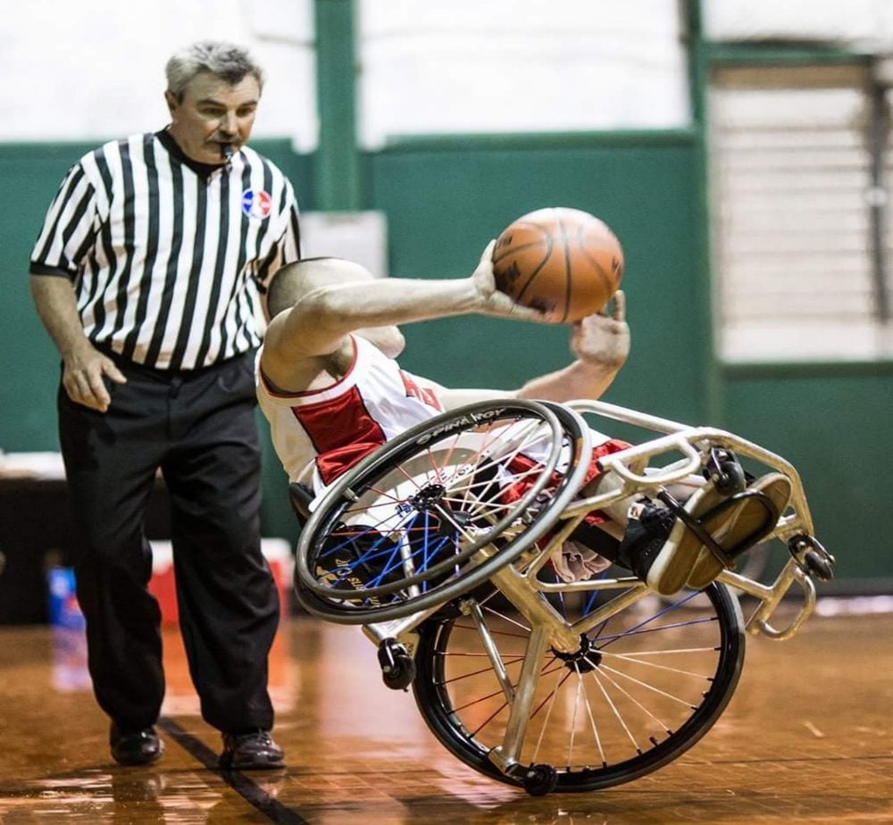 A New York Rolling Fury player reaching for the ball mid-play in a wheelchair basketball game as a referee looks on