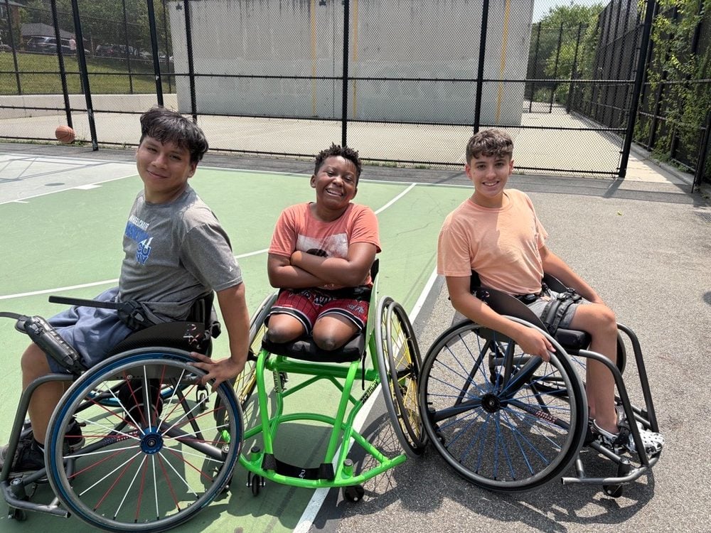 Three young Rolling Fury athletes in wheelchairs smiling together at an outdoor practice court